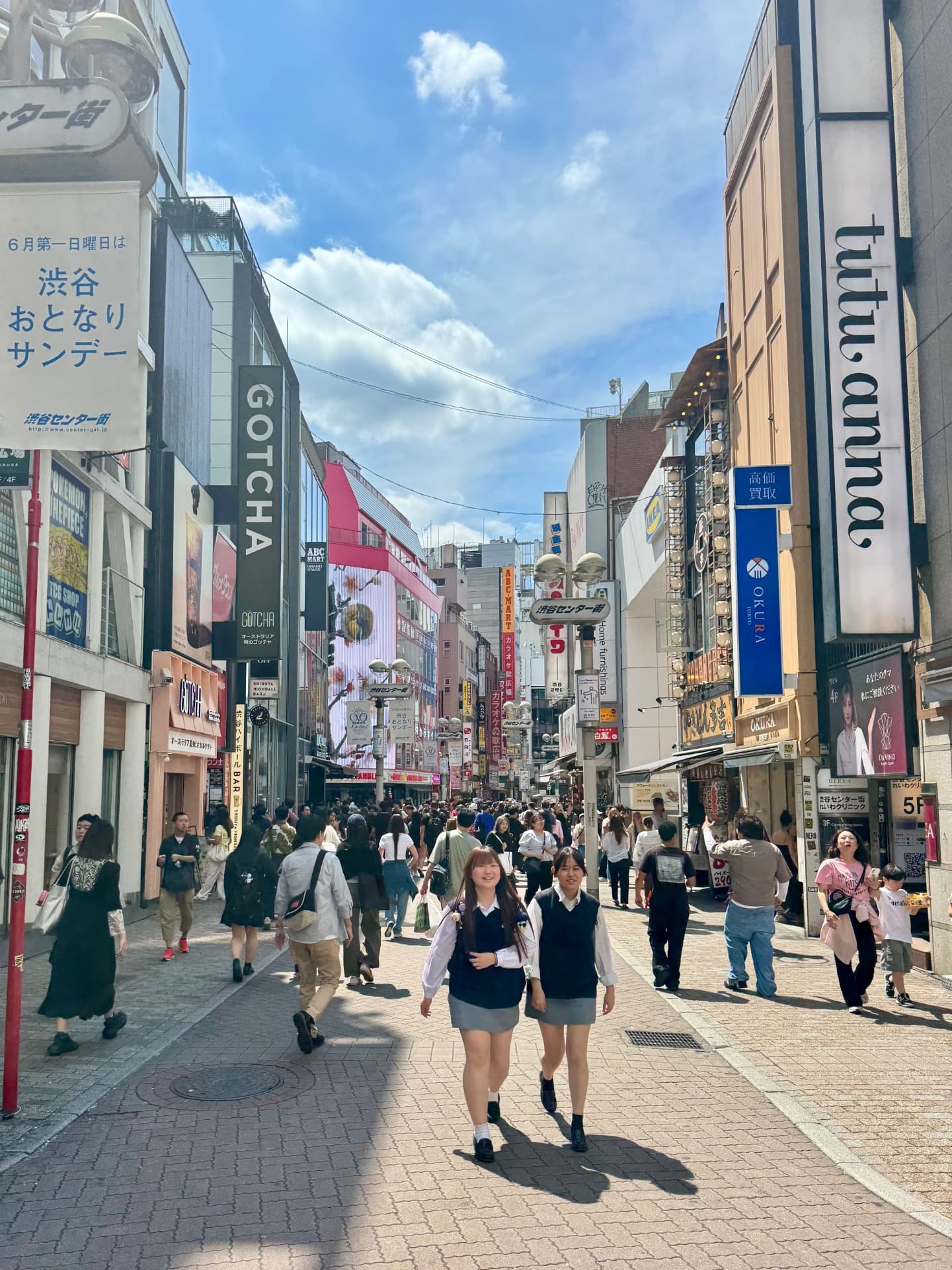 Busy pedestrian shopping street in Shibuya, Tokyo with crowds of people, colorful storefronts, and blue sky overhead.