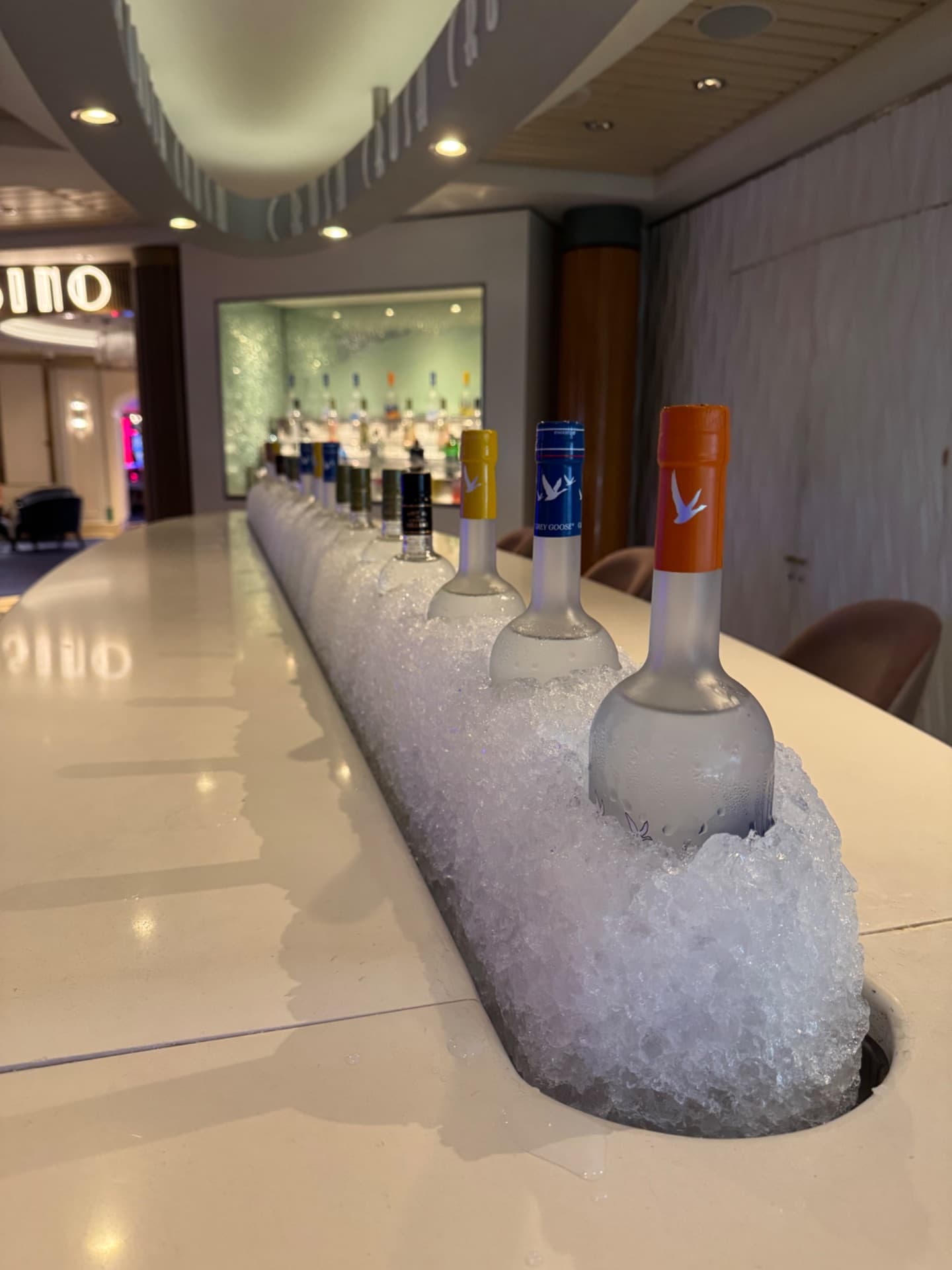 Wine bottles chilled in ice along a bar counter in an elegant cruise ship lounge with modern lighting and seating.