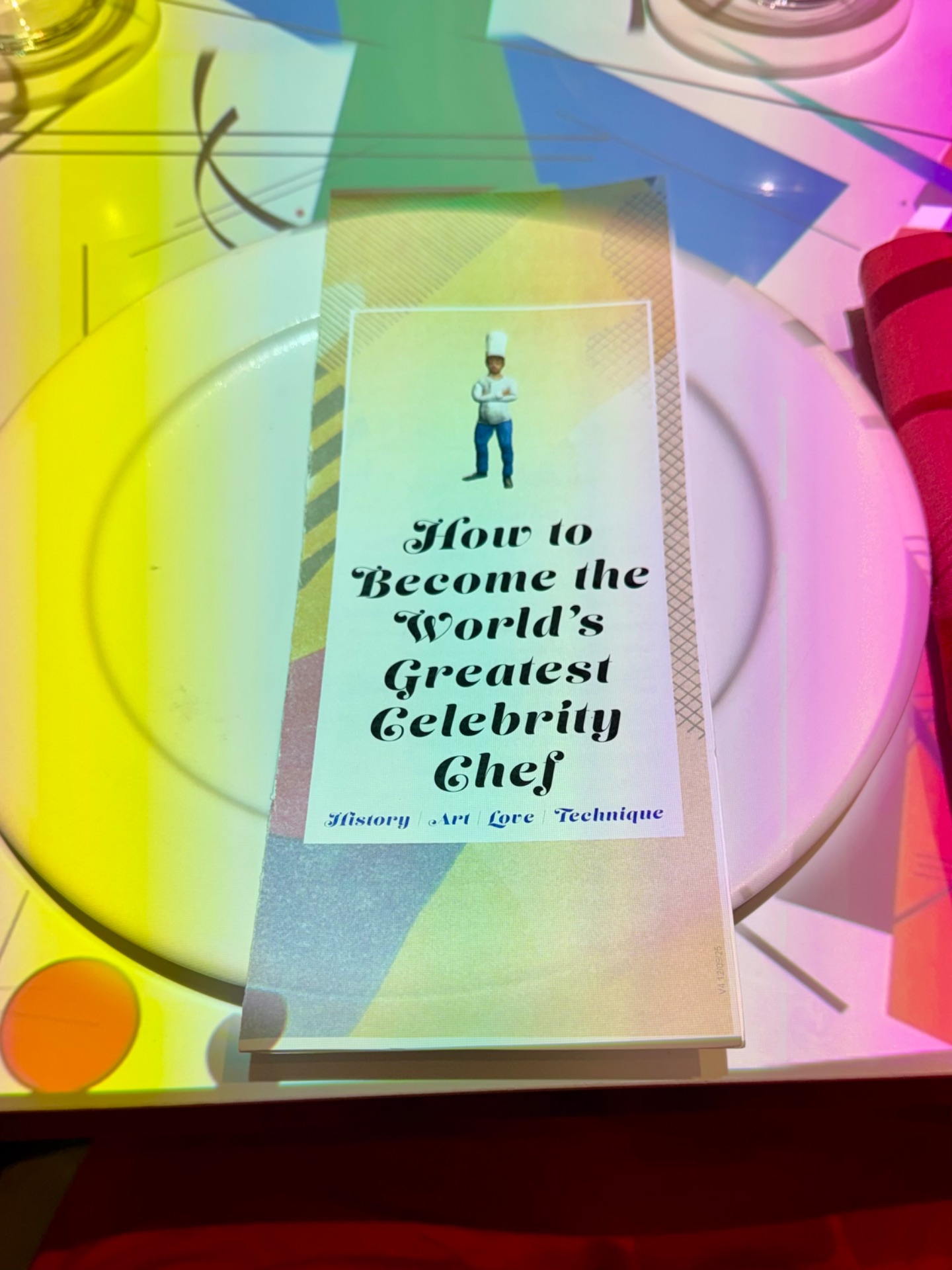 A colorful cruise ship menu booklet titled "How to Become the World's Greatest Celebrity Chef" surrounded by vibrant tableware and plates.
