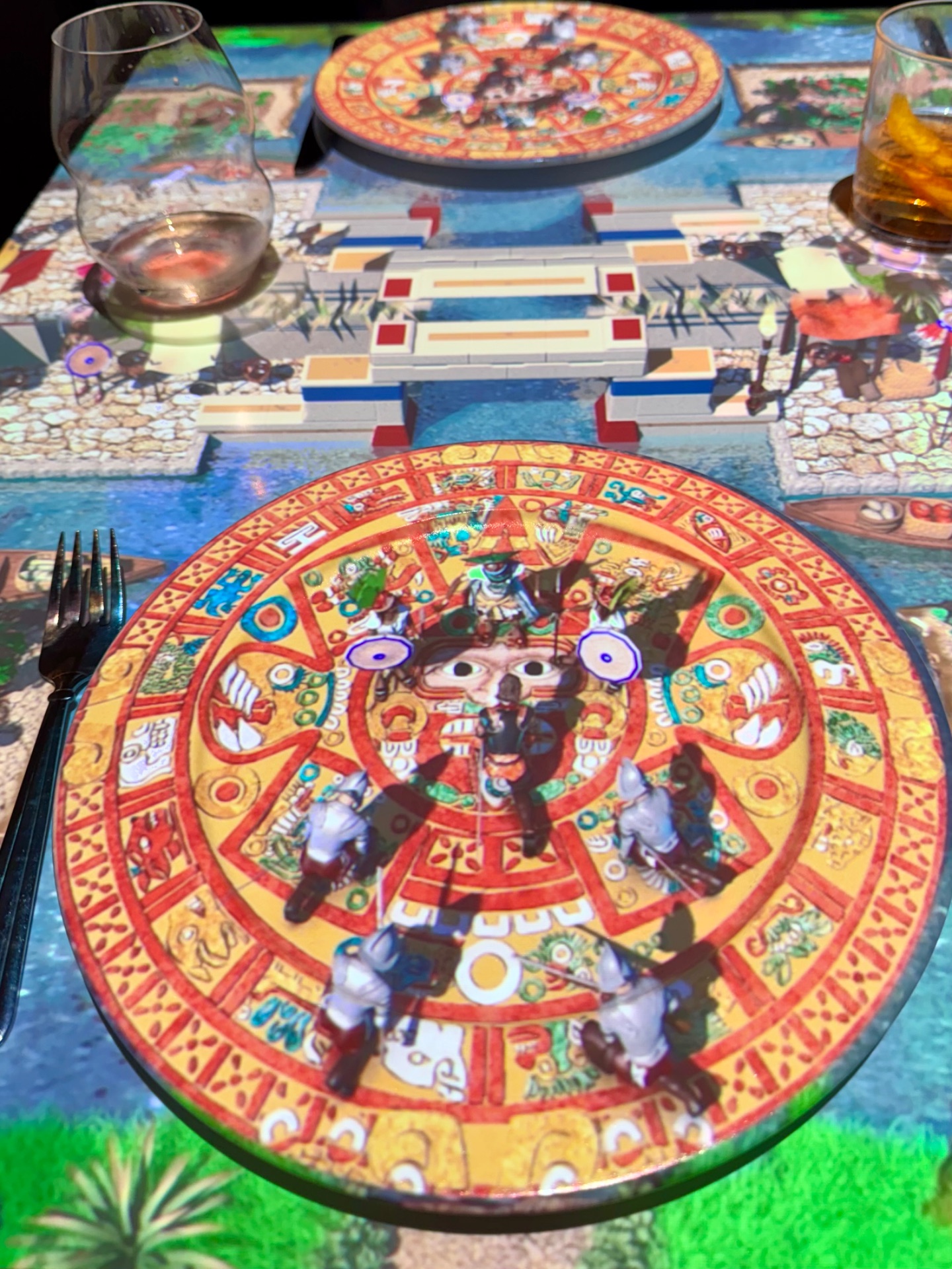 Colorful Mayan-themed dinner plates at an elegant cruise ship dining table with decorative placemat and glassware.