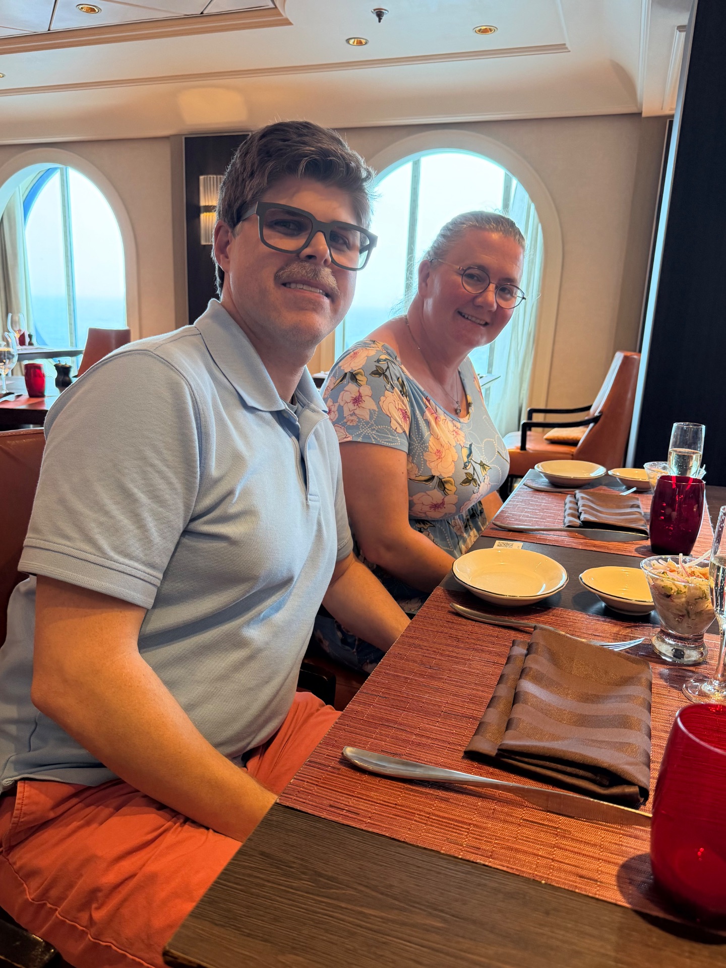 Two passengers dining together in a cruise ship's formal dining room with ocean views through porthole windows.