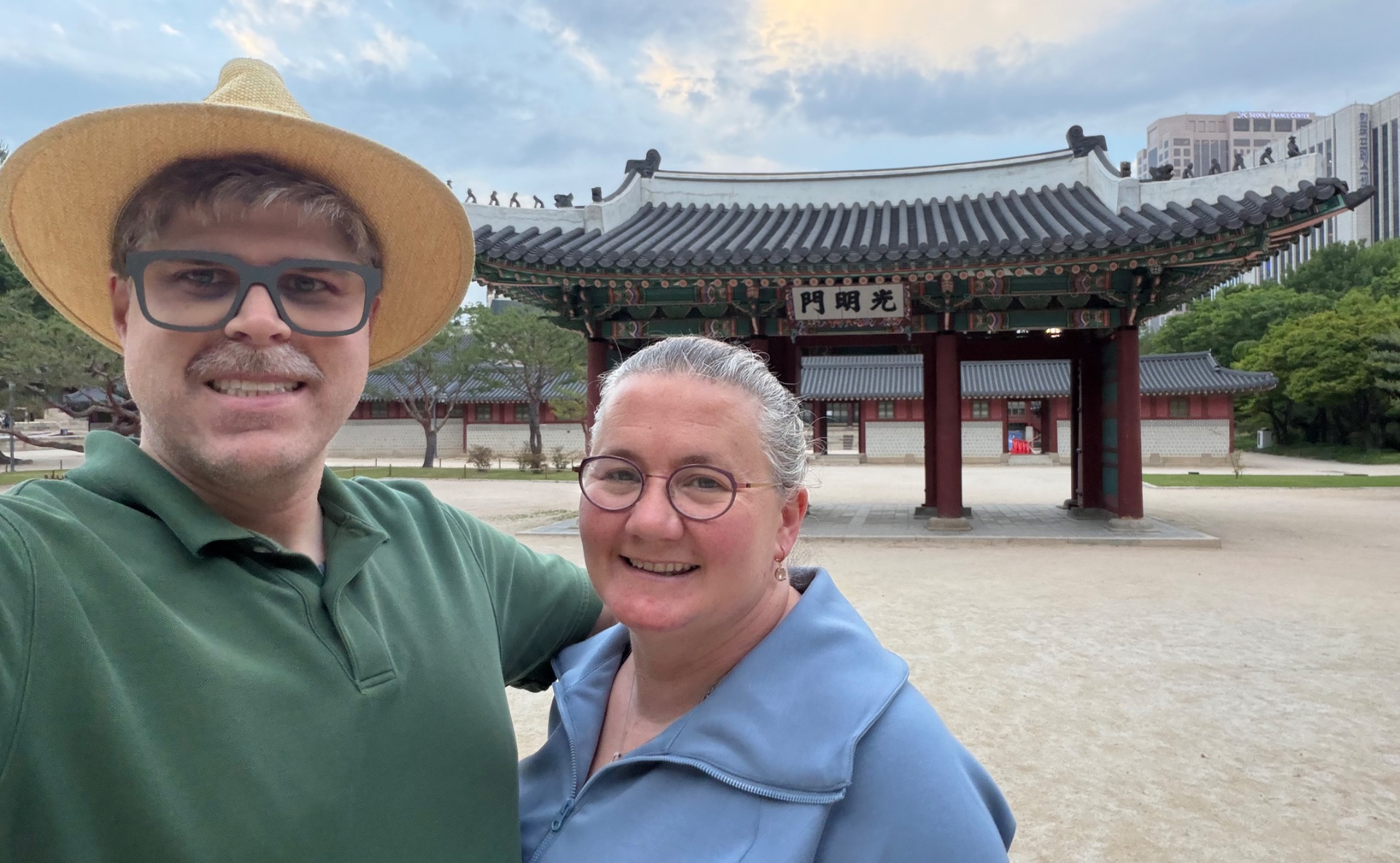 Two tourists smile at a traditional Korean temple with ornate curved roof and red wooden gates in the background.
