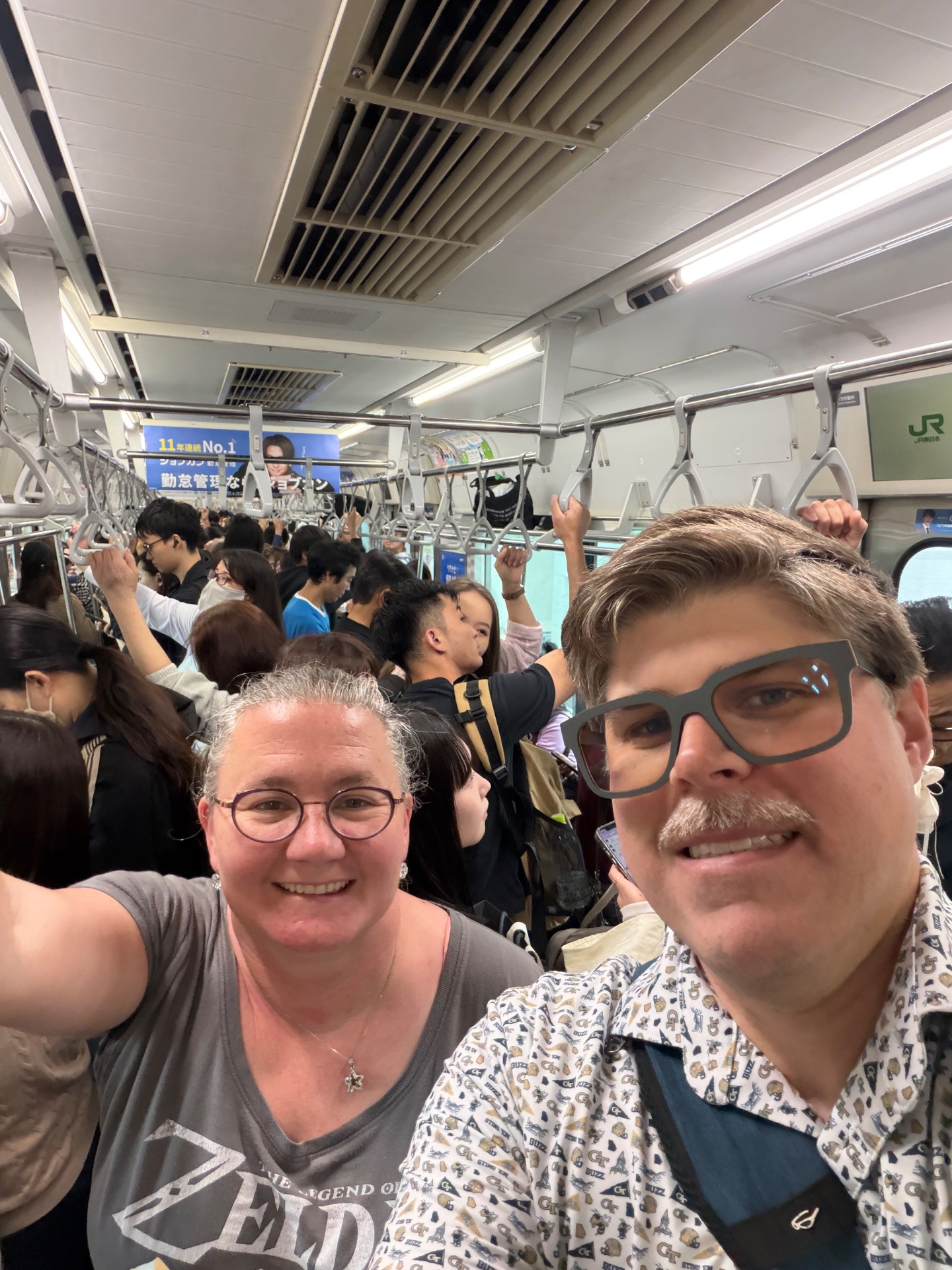 Two tourists smile for a selfie on a crowded JR train in Japan during daytime travel.