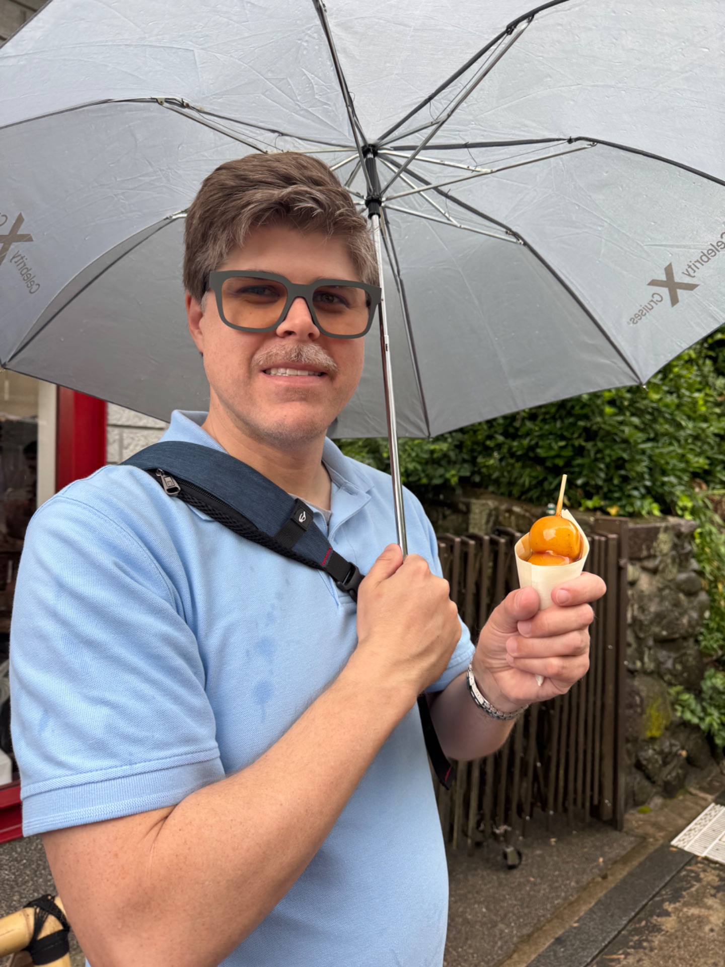 Man holding umbrella and dessert in rainy outdoor setting, wearing glasses and blue polo shirt
