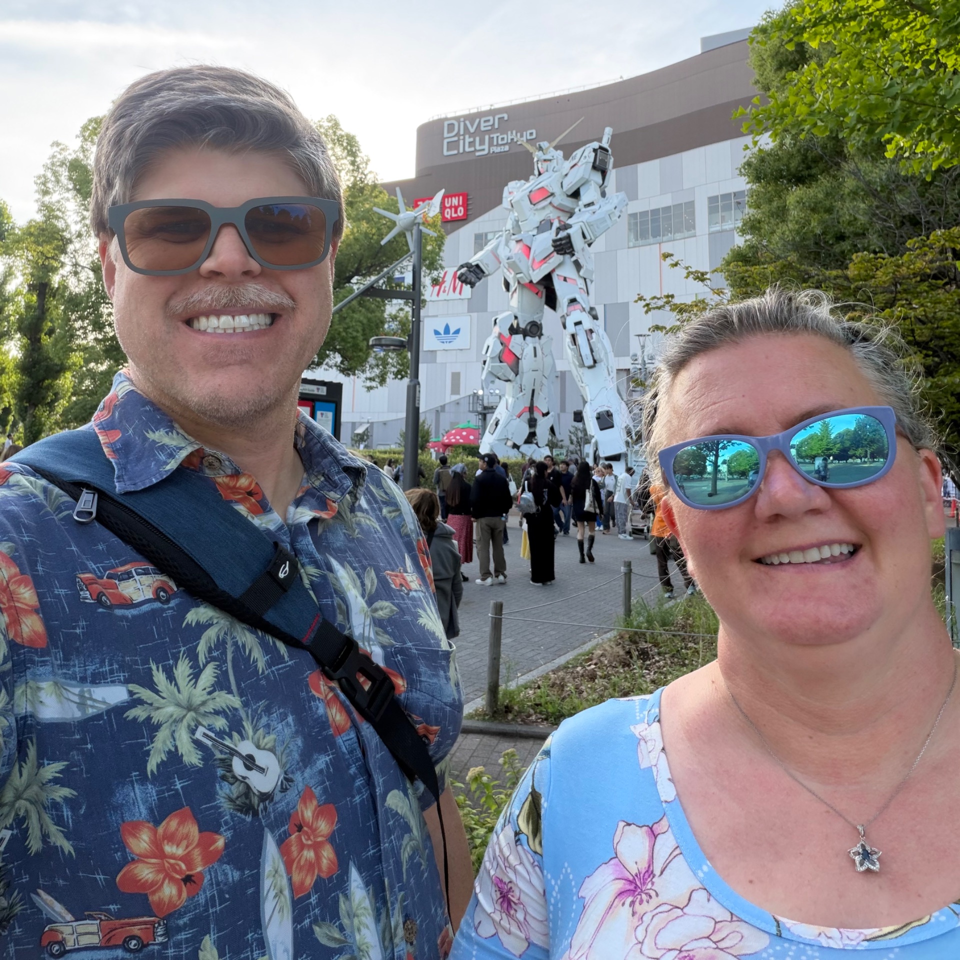 Two smiling visitors pose in front of a giant white and red Gundam statue at Diver City Tokyo Plaza shopping complex.