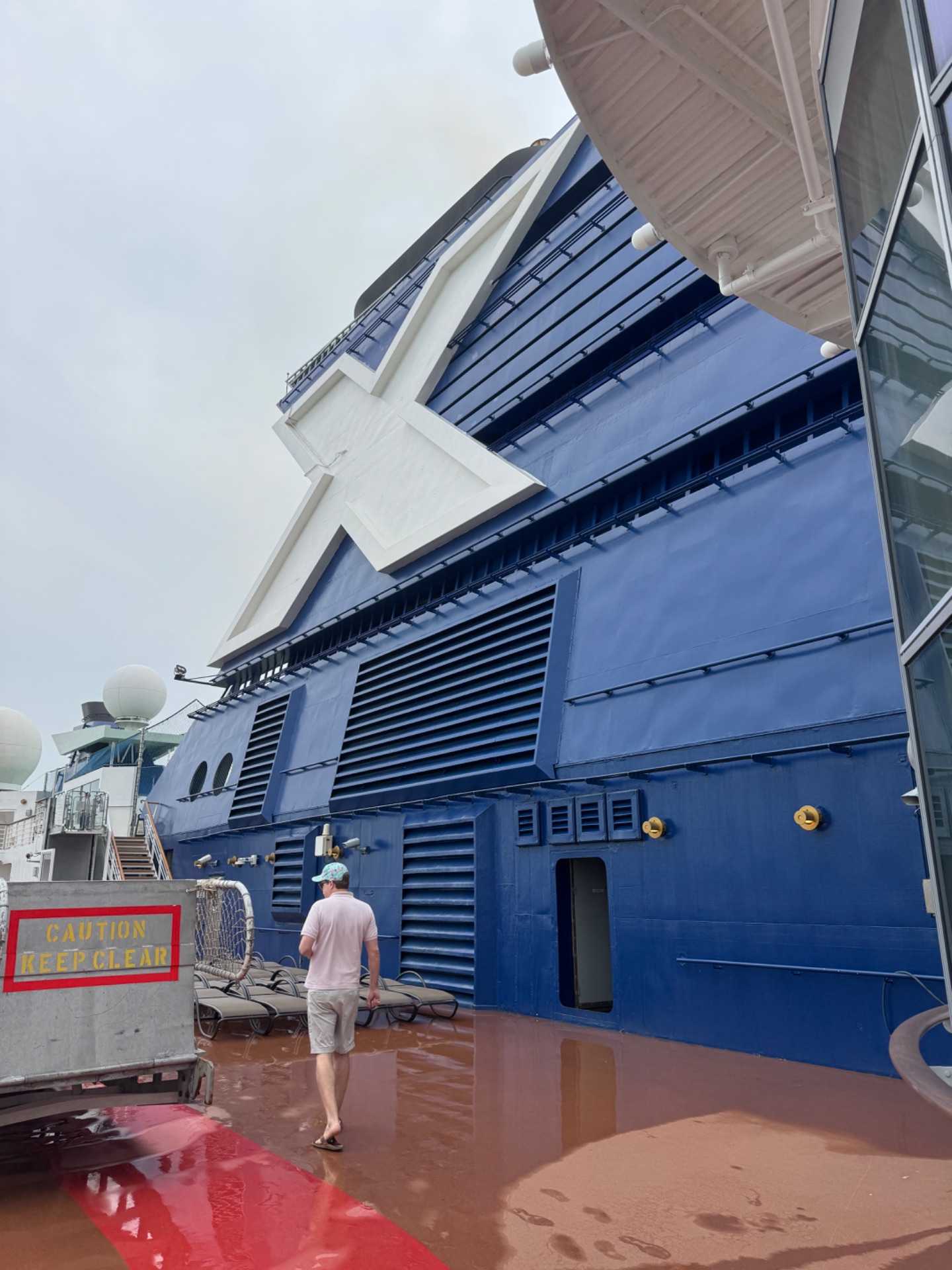 Person walking on wet deck beside large blue cruise ship with extended awning and white superstructure.
