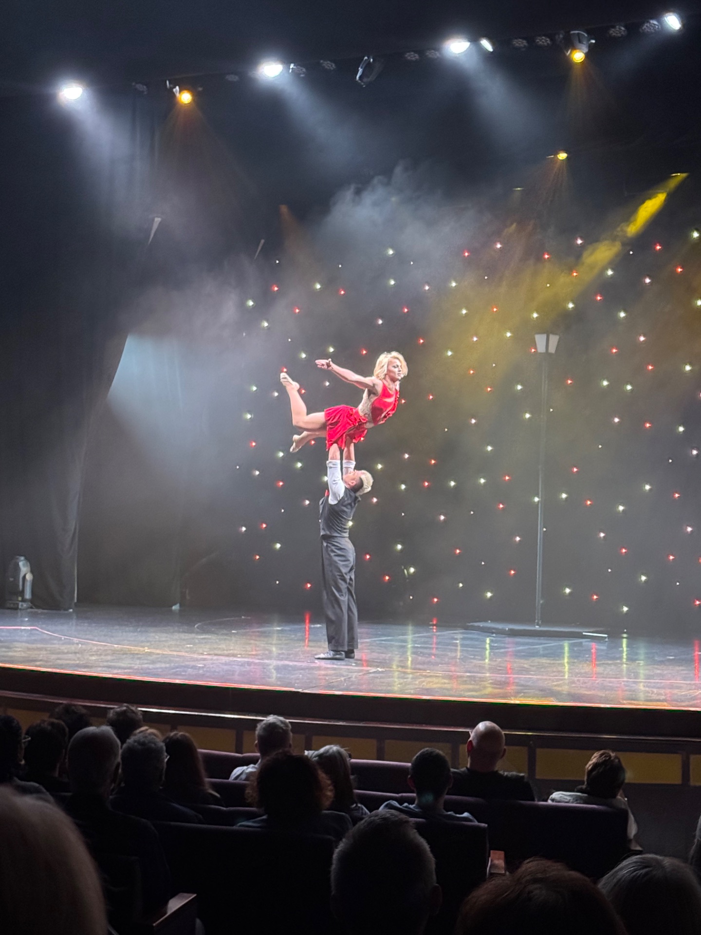 Acrobat in pink costume lifted high above stage during circus performance with theatrical lighting and audience in foreground.