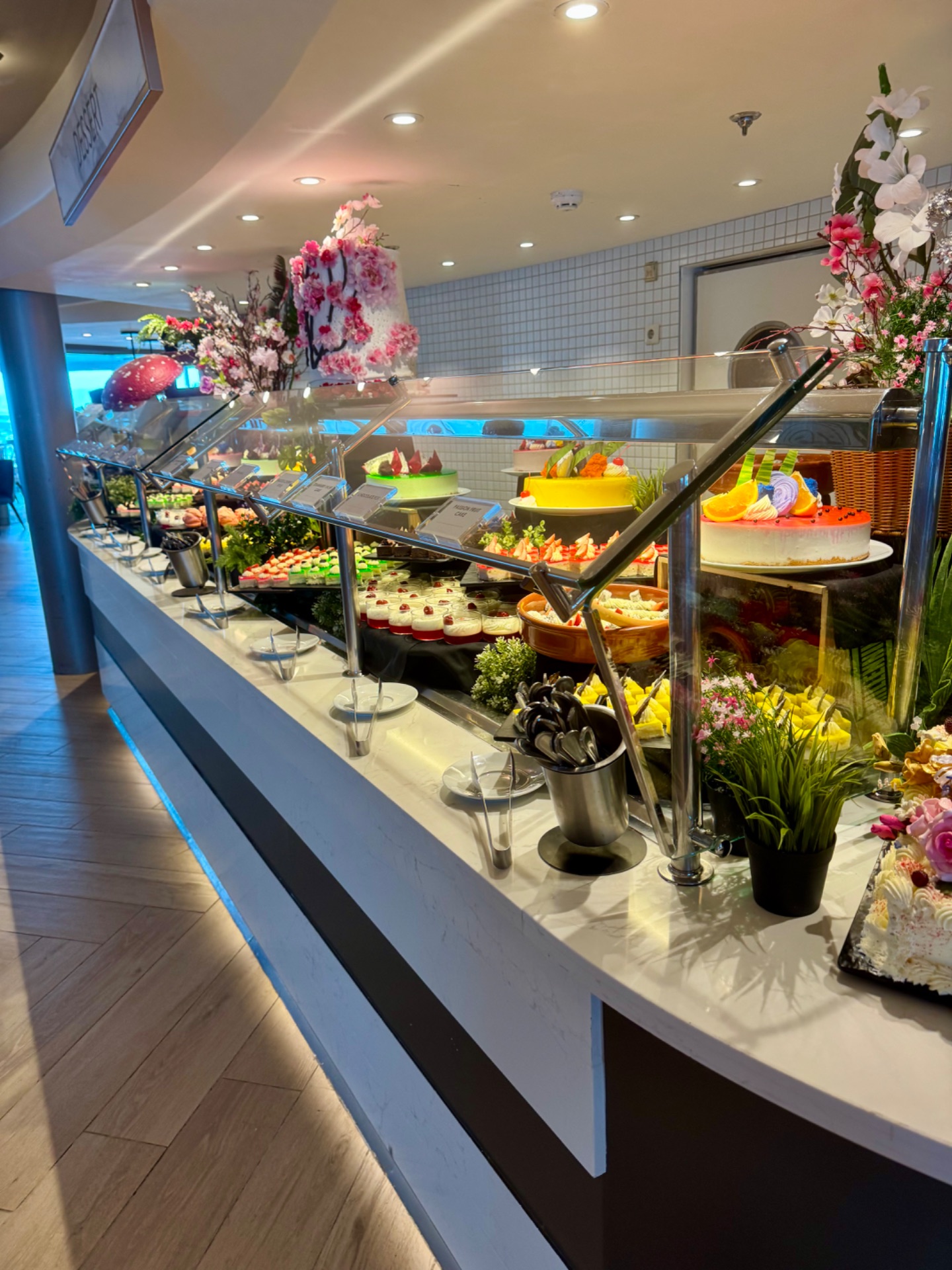 Elegant cruise ship pastry display counter with colorful cakes, desserts, and floral decorations under ambient lighting.