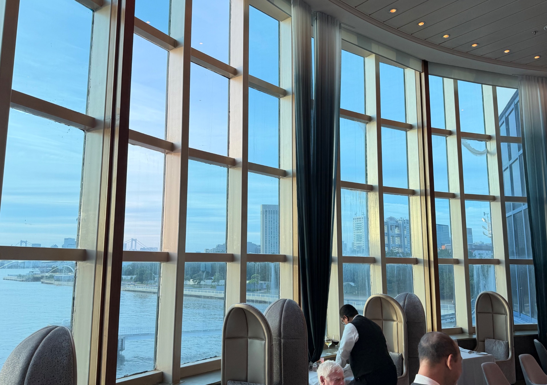 Passengers enjoying waterfront views from a cruise ship dining room with floor-to-ceiling windows overlooking a city skyline and bridges.