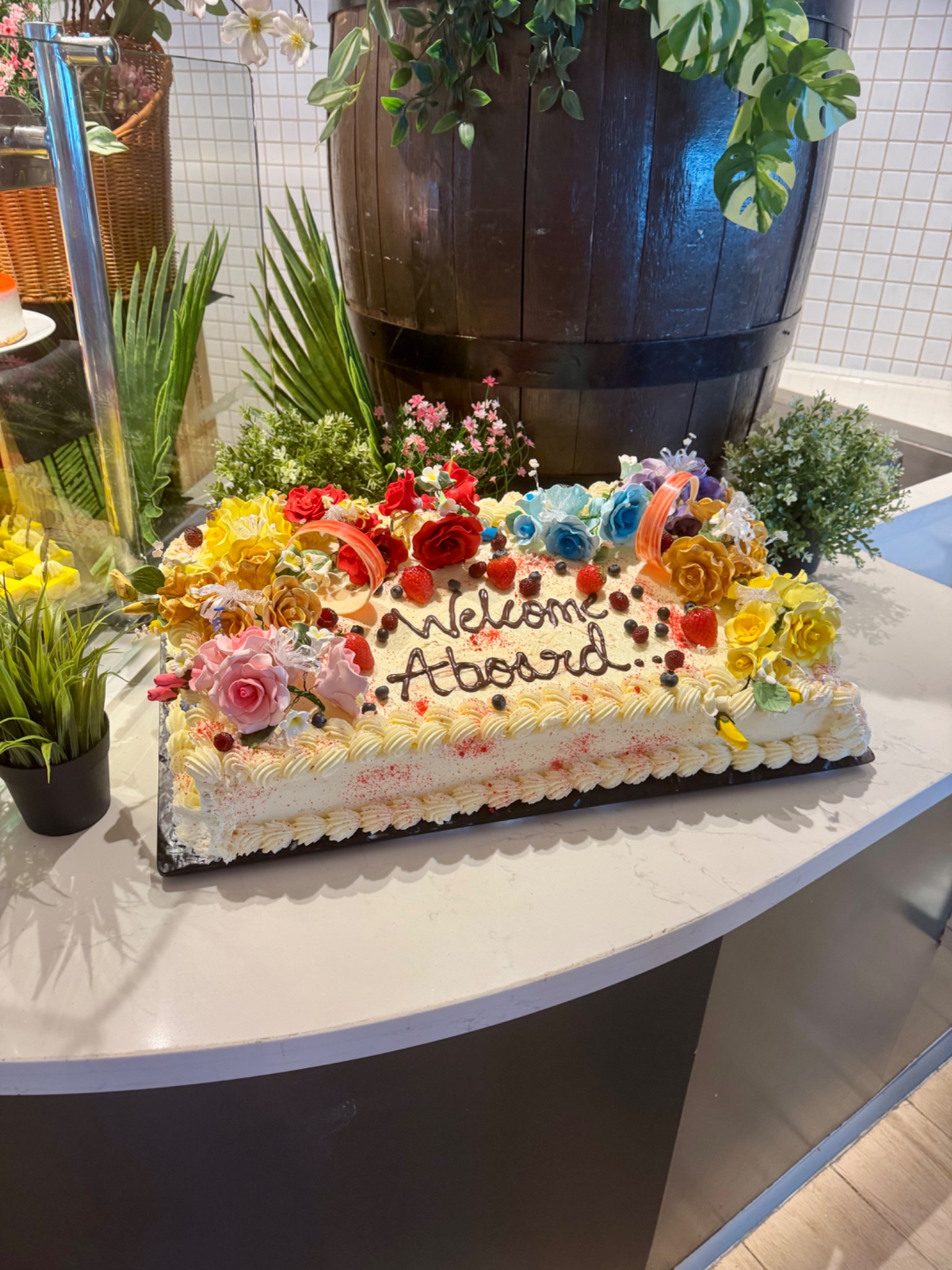 Elaborately decorated "Welcome Aboard" cake with colorful sugar flowers and fresh berries on a cruise ship counter.