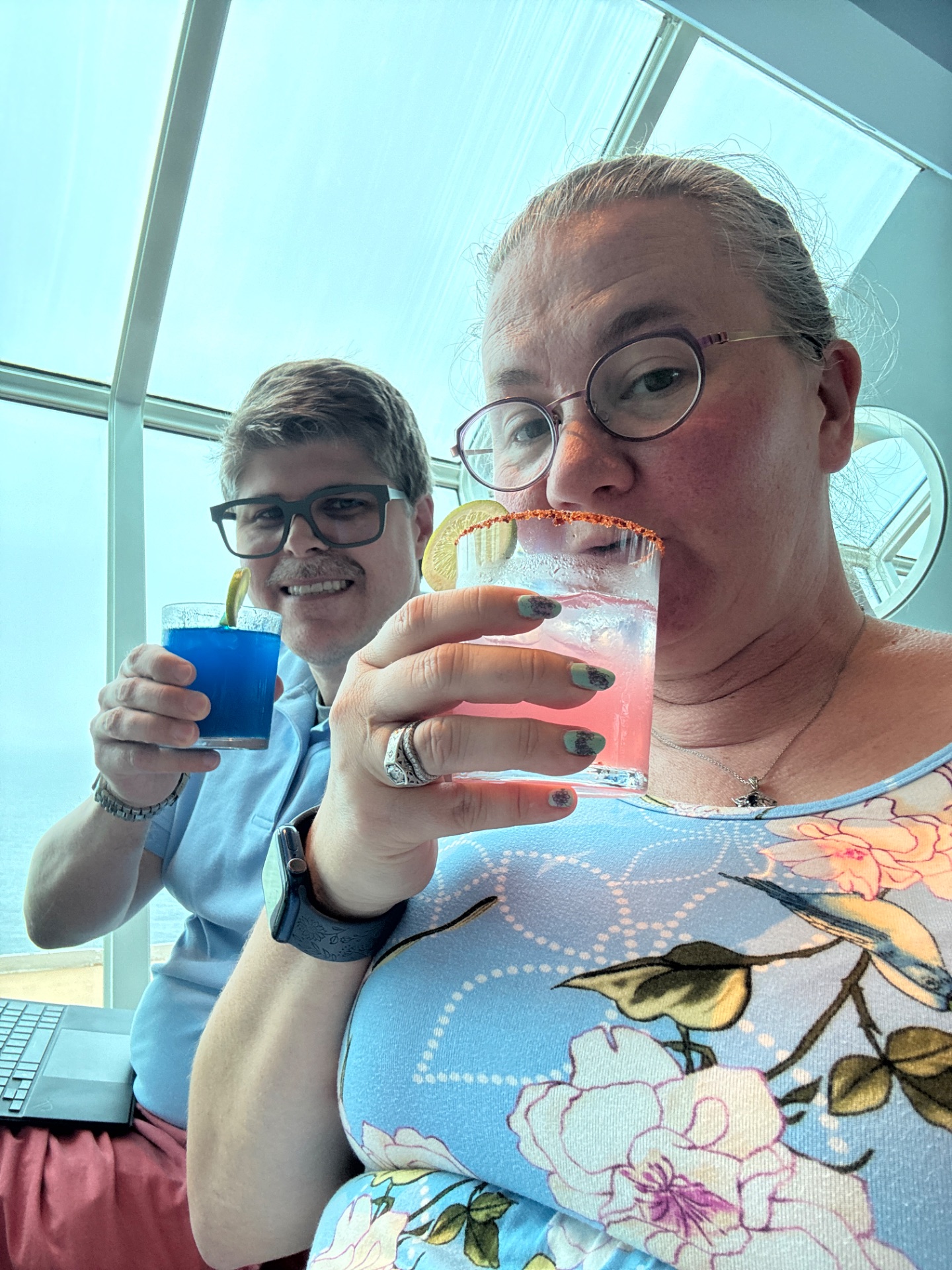 Two people enjoying colorful cocktails by large windows on a cruise ship with ocean views.
