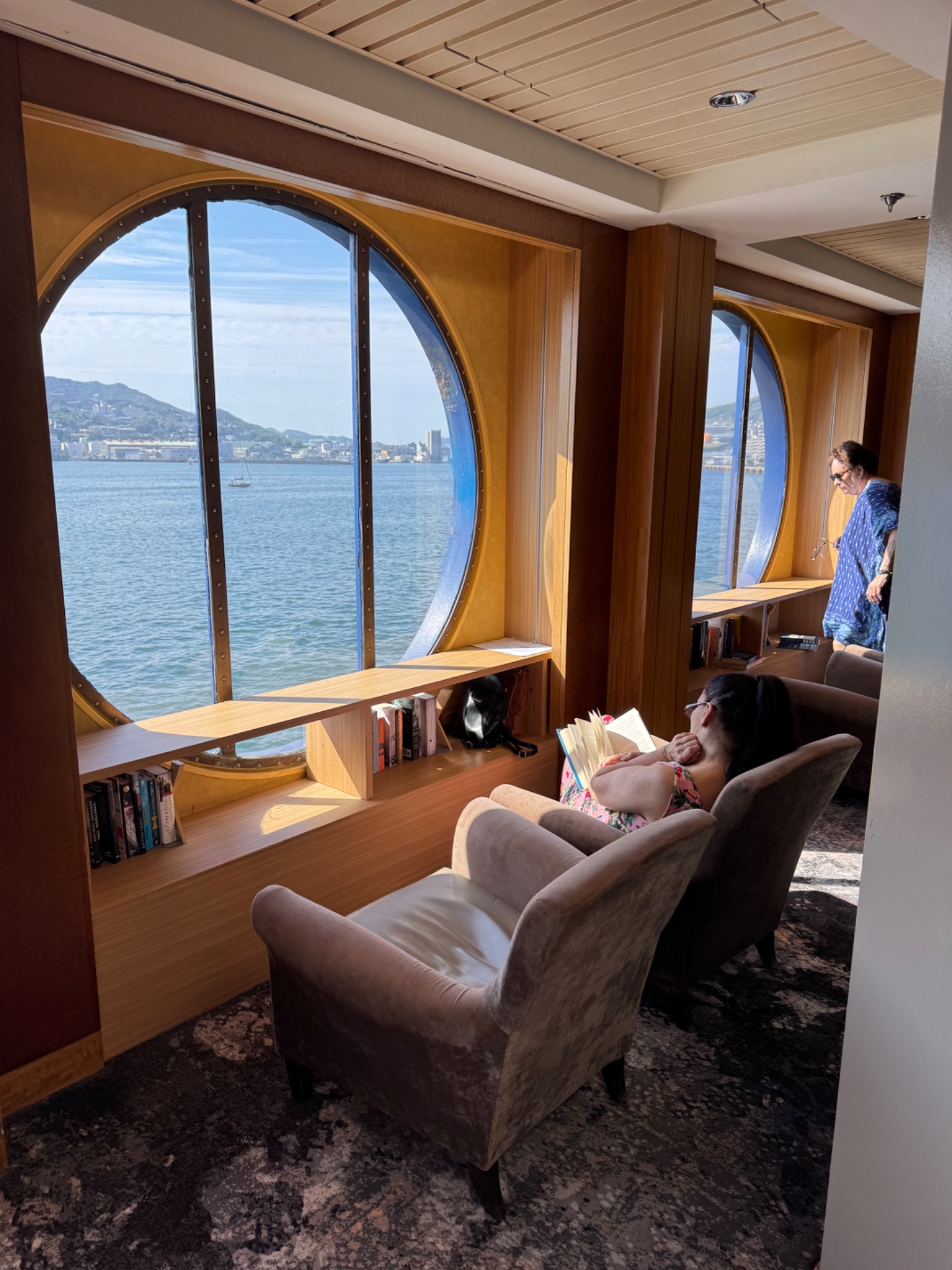 Passenger relaxing in a cruise ship cabin with circular porthole window overlooking coastal waters and hillside buildings.
