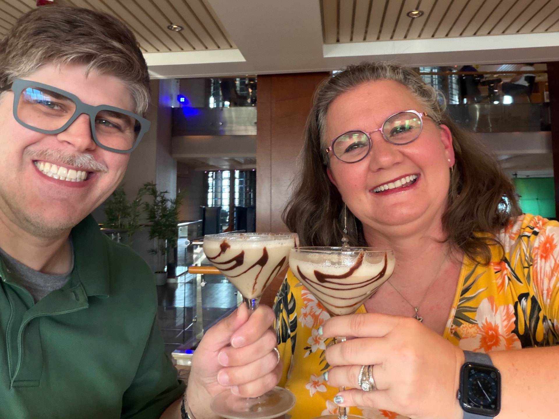 Two people smiling at a cruise ship bar while holding decorative cocktails with chocolate drizzle designs.