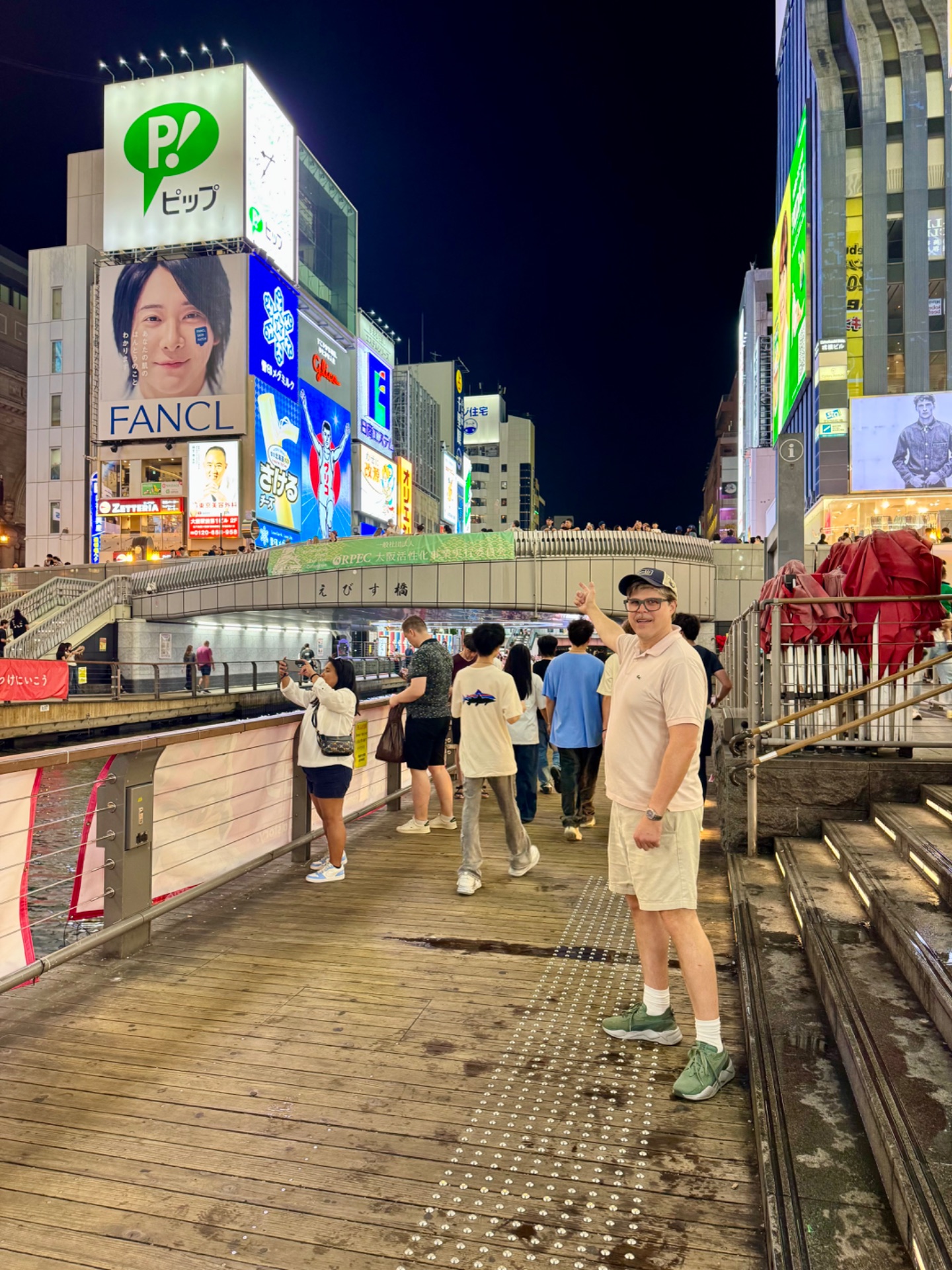 Man in casual attire posing on wooden boardwalk at night in Japanese shopping district with illuminated signs and buildings.