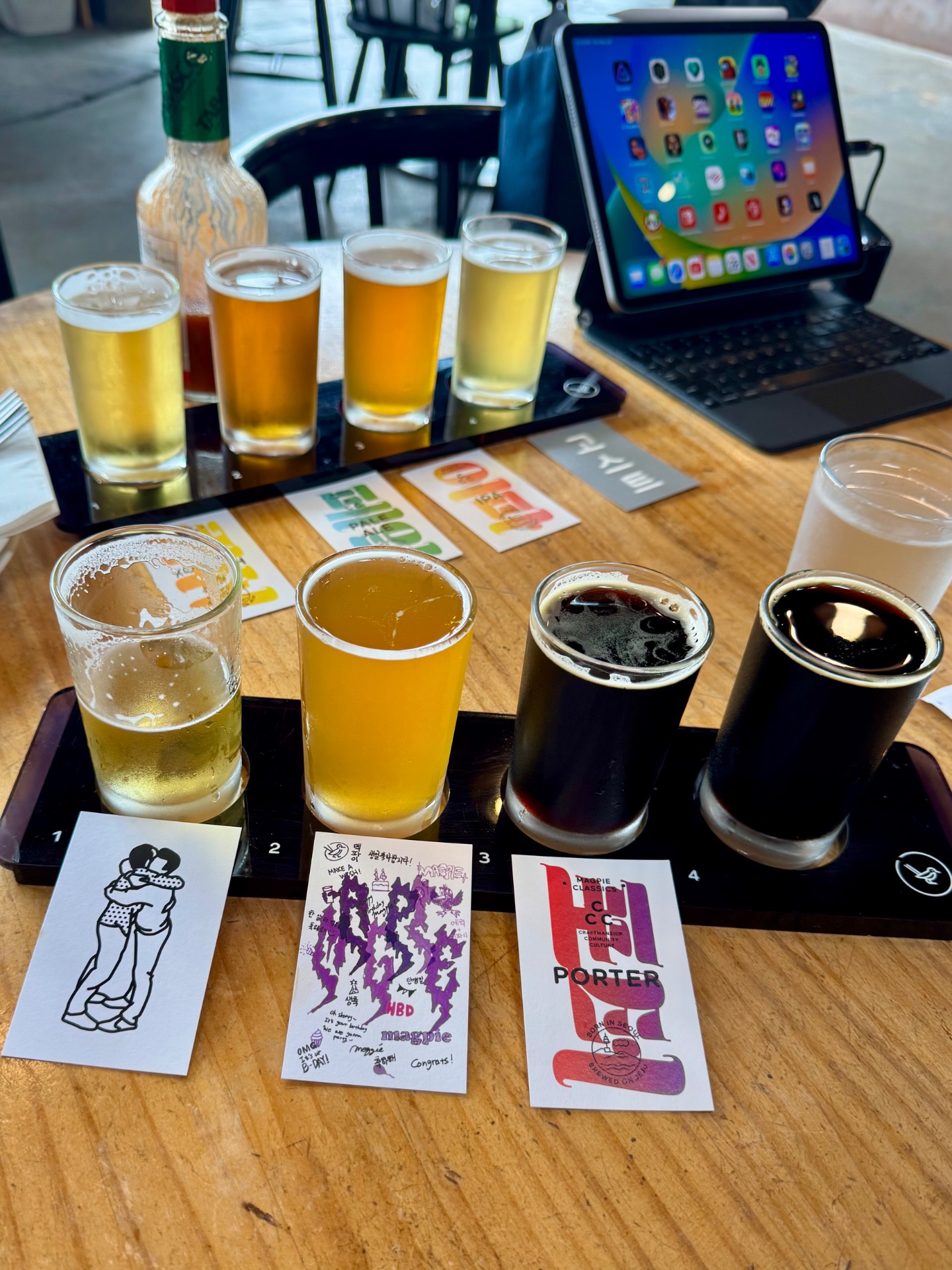 Beer flight tasting at a brewery with four labeled samples, iPad, and bottle on wooden table.