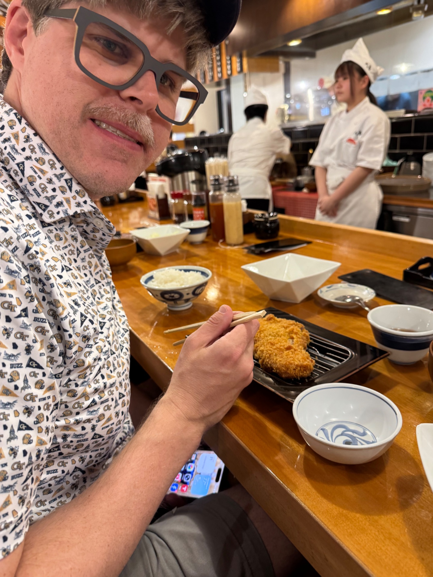Man wearing glasses at a sushi bar counter holding a fried item, with chefs working in background and condiments on wooden counter.