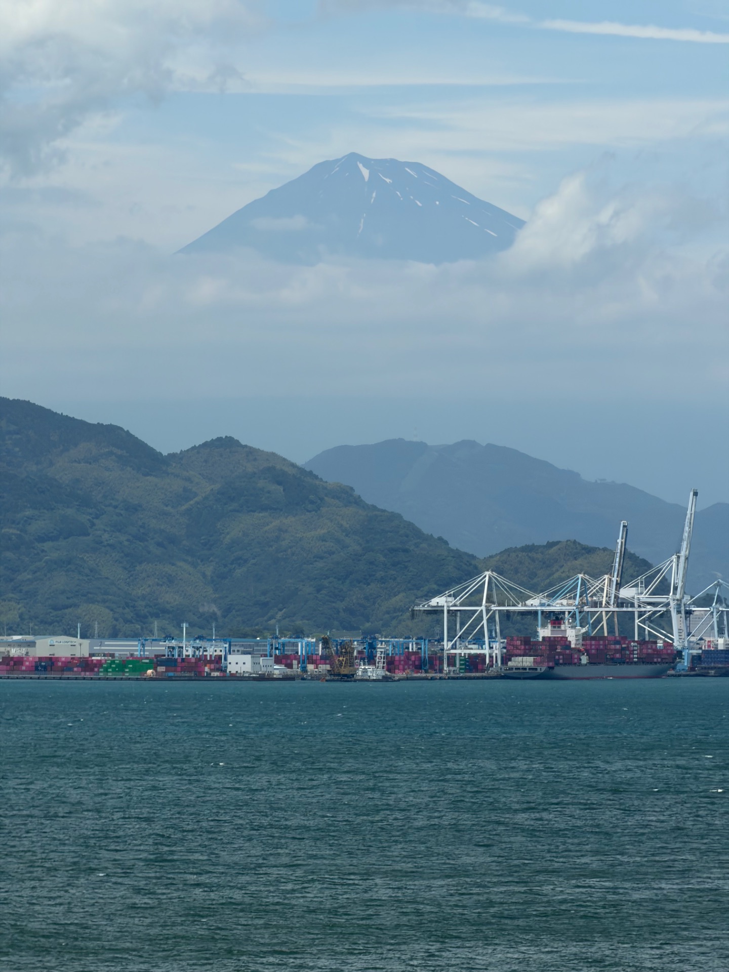 Mount Fuji rises above clouds behind a busy container port with cargo ships and cranes along the Japanese coast.