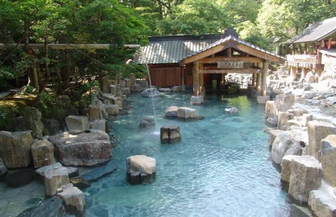Traditional Japanese hot spring bathhouse surrounded by turquoise water and natural stone rocks in a forest setting.