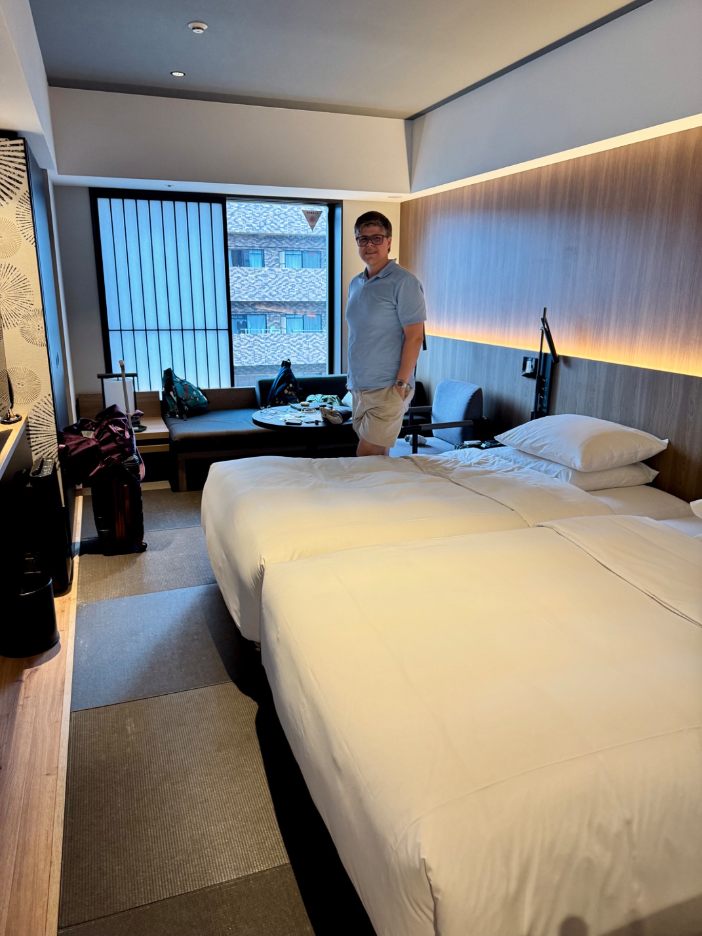 Man standing in modern hotel room with two beds, sofa, and ocean view from floor-to-ceiling windows.