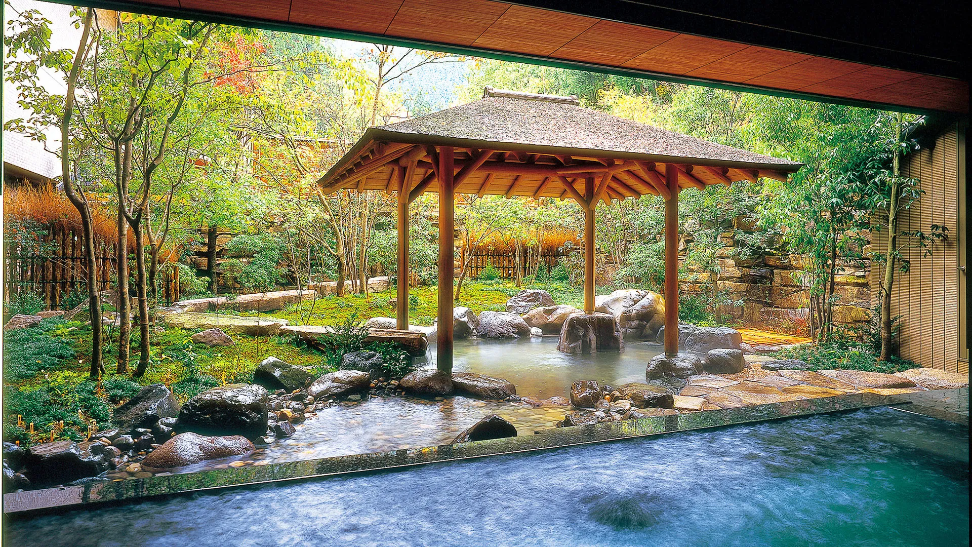 Japanese-style wooden pavilion overlooking a natural hot spring with rocks and lush green forest surroundings.