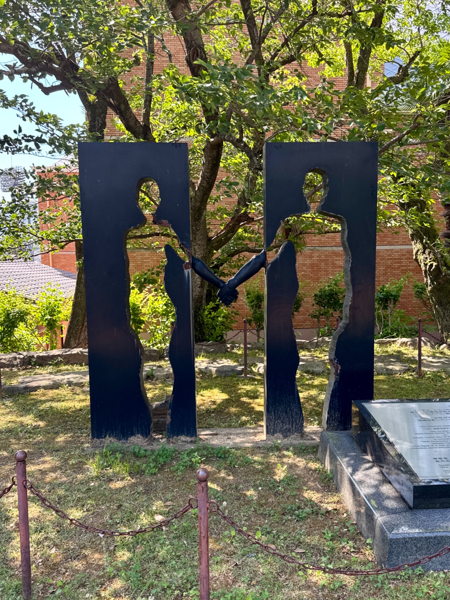 Modern black metal sculpture of two figures holding hands in a sunlit garden with brick buildings and trees in background.