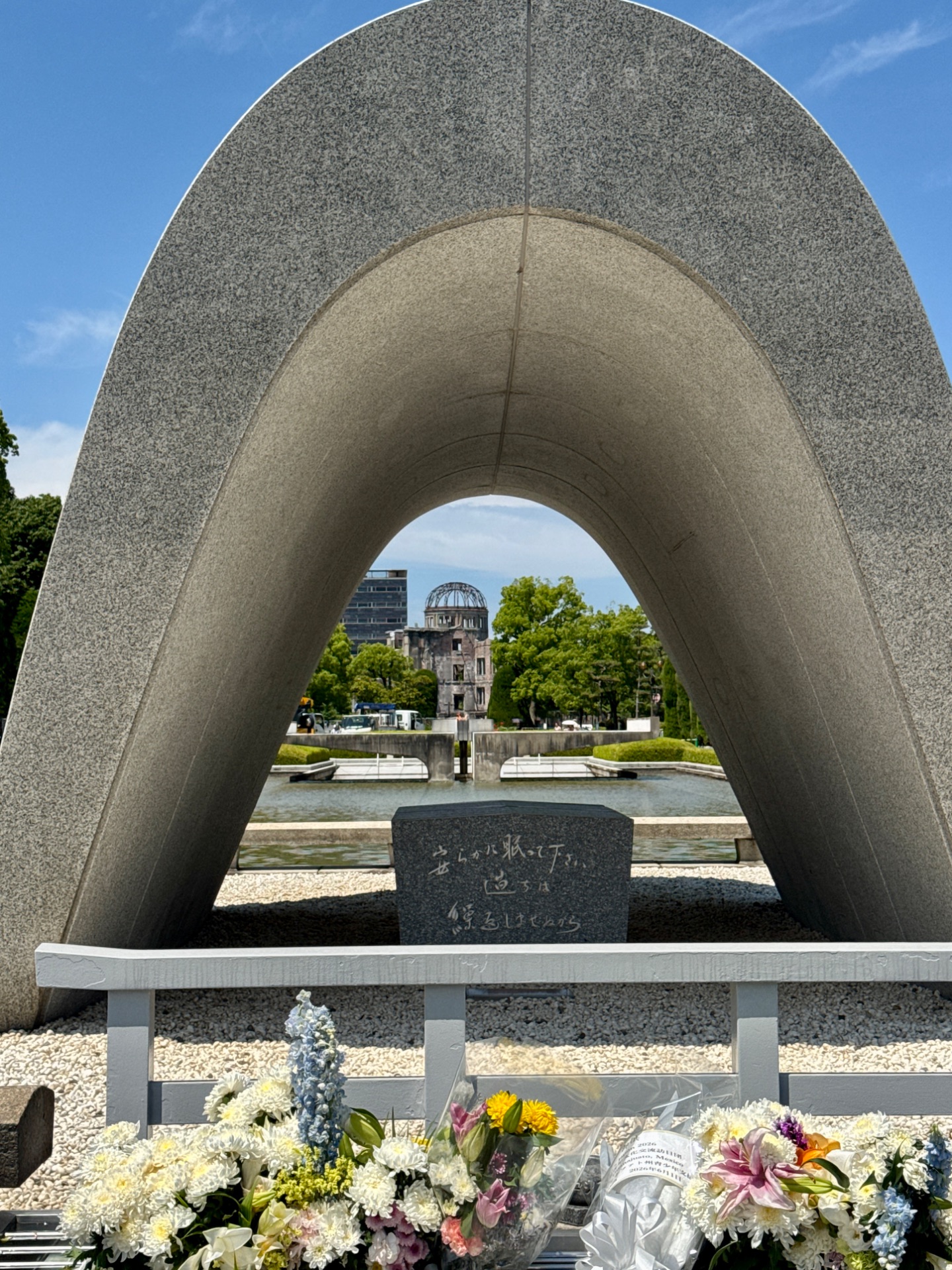 Cenotaph memorial with arched stone frame overlooking Hiroshima's A-Bomb Dome across the river, surrounded by flowers.