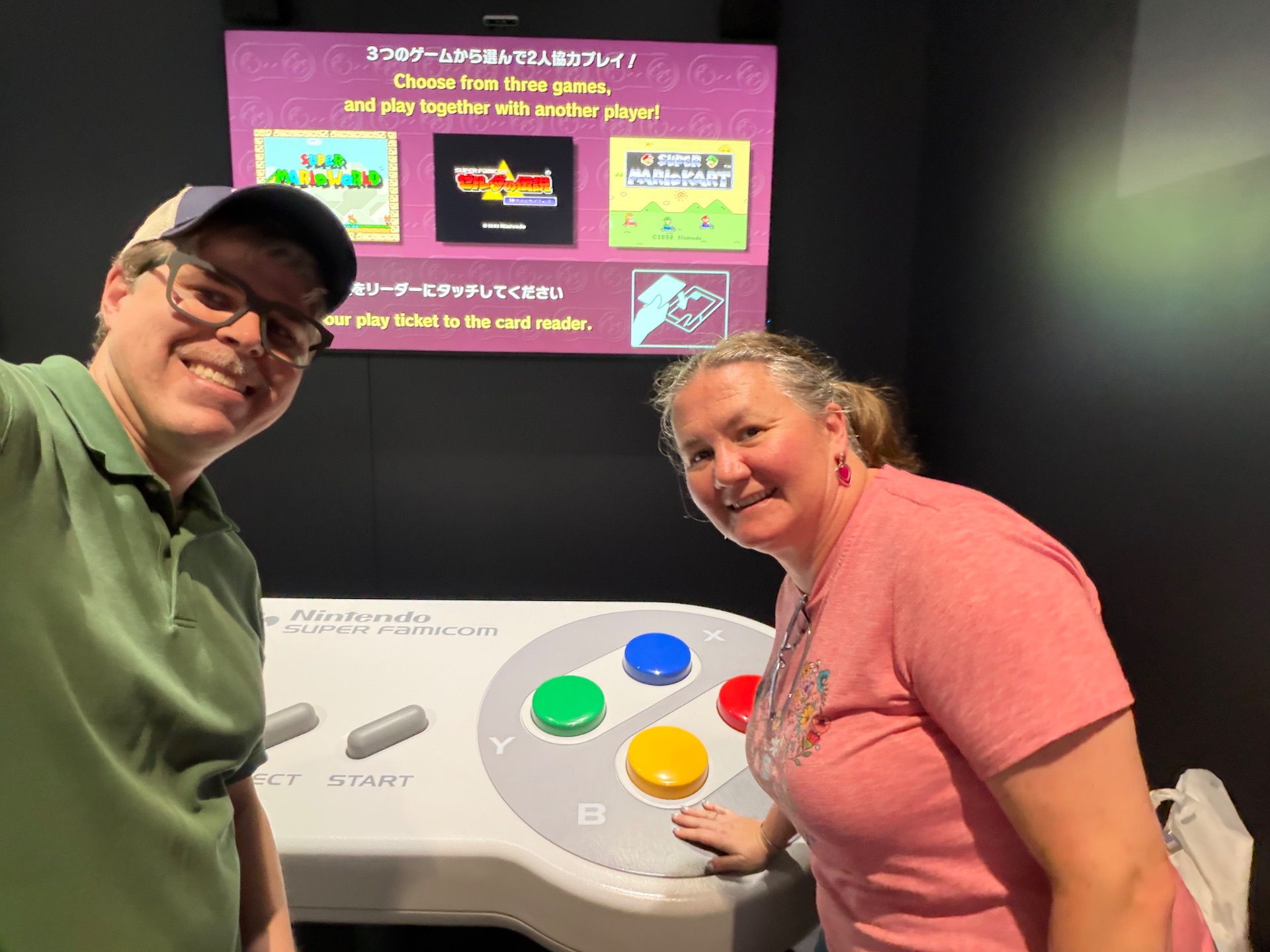 Two people posing at a Nintendo Super Famicom arcade game station with colorful buttons and game selection screen displayed above.