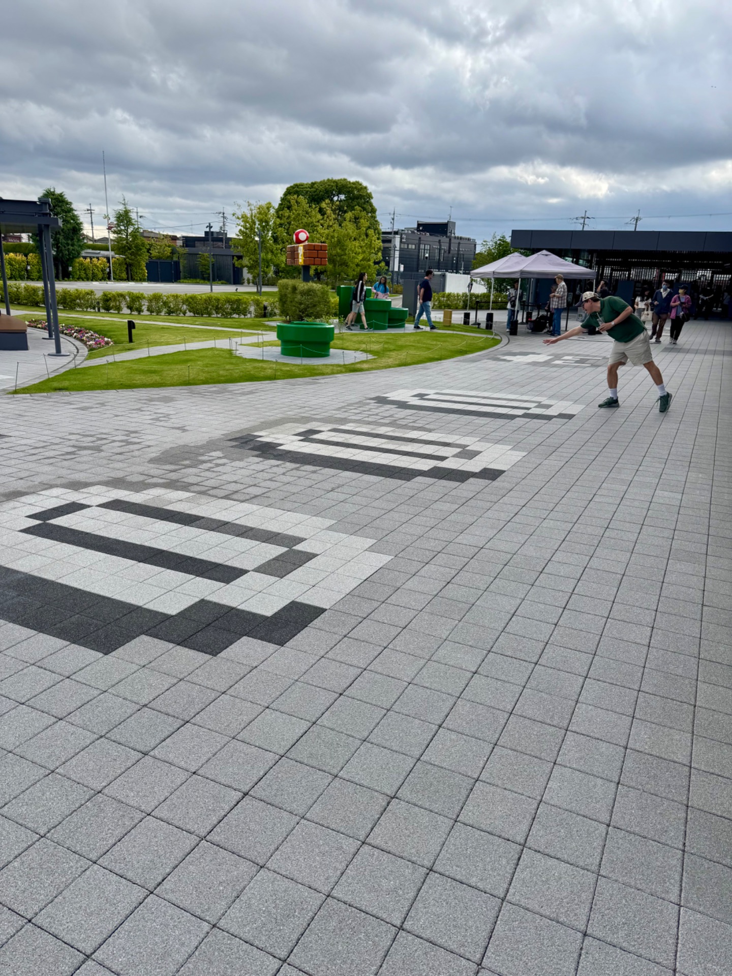 Modern public plaza with paved grounds, green spaces, and visitors enjoying an outdoor event under cloudy skies.